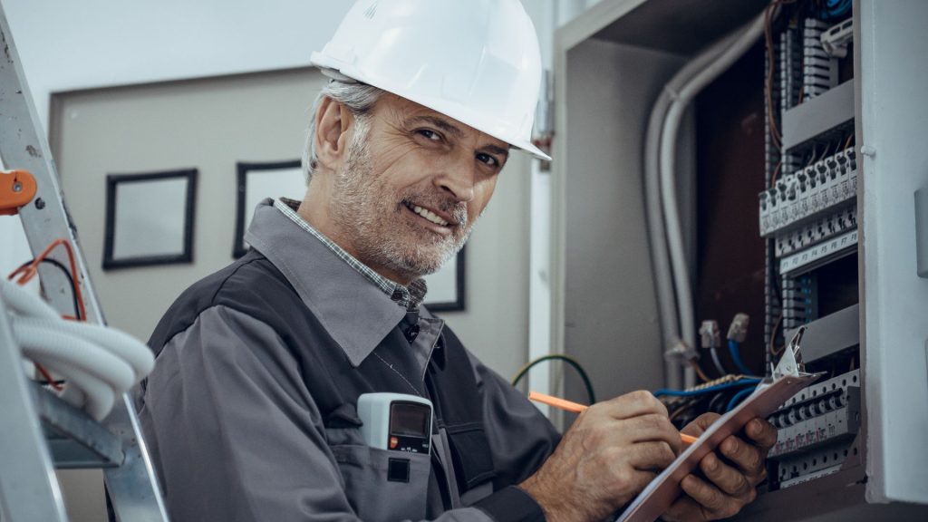 Mann mit Sicherheitshelm steht vor einem Sicherungskasten. Er hat ein elektrisches Prüfgerät in der Tasche und dokumentiert eine elektrische Prüfung.